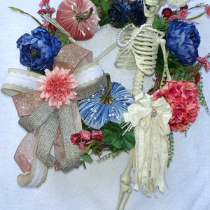 May include: A Halloween wreath with a skeleton, blue and pink velvet pumpkins, a pink and brown bow, and a white flower.
