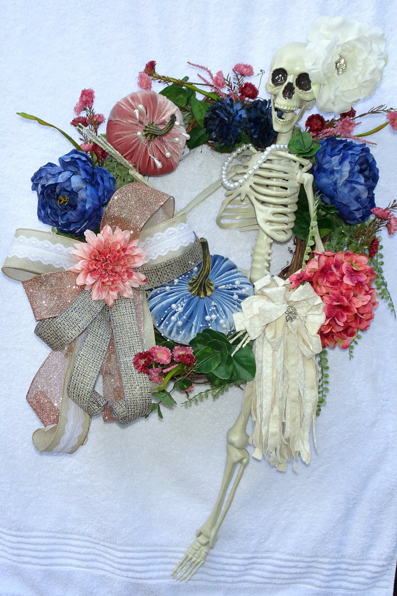May include: A Halloween wreath with a skeleton, blue and pink velvet pumpkins, a pink and brown bow, and a white flower.