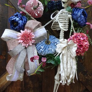 May include: A Halloween wreath with a skeleton, pumpkins, and flowers in shades of pink, blue, and white. The wreath is decorated with a pink and silver bow and a white fabric bow.