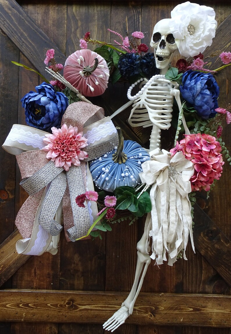 May include: A Halloween wreath with a skeleton, pumpkins, and flowers in shades of pink, blue, and white. The wreath is decorated with a pink and silver bow and a white fabric bow.