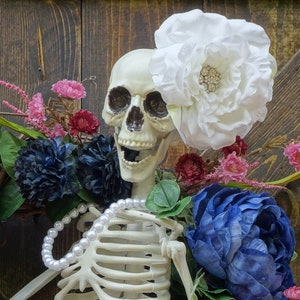 May include: A white skeleton with a pearl necklace and a large white flower on its head. The skeleton is surrounded by pink, blue, and green flowers.