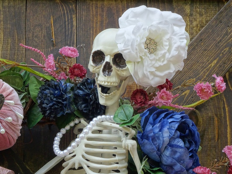 May include: A white skeleton with a pearl necklace and a large white flower on its head. The skeleton is surrounded by pink, blue, and green flowers.