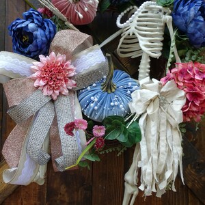 May include: A blue and white velvet pumpkin with a pink and white bow, a white and pink flower, and a white fabric bow with a silver embellishment. A skeleton is in the background with blue and pink flowers.