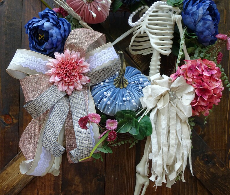 May include: A blue and white velvet pumpkin with a pink and white bow, a white and pink flower, and a white fabric bow with a silver embellishment. A skeleton is in the background with blue and pink flowers.
