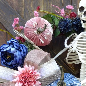 May include: A pink velvet pumpkin, blue flowers, and a white skeleton with a pearl necklace are arranged on a wooden background. The skeleton is wearing a white pearl necklace.
