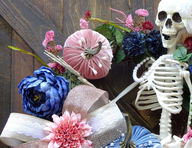 May include: A pink velvet pumpkin, blue flowers, and a white skeleton with a pearl necklace are arranged on a wooden background. The skeleton is wearing a white pearl necklace.