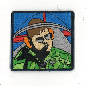Puede incluir: Un parche bordado de dibujos animados de un piloto con un traje de vuelo verde, gafas de sol y auriculares. El piloto está delante de un avión azul y rojo. El texto "caution pilot" está en la esquina inferior derecha del parche.