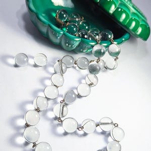 May include: A clear glass beaded necklace with a green malachite shell box. The necklace is made of large, round beads and is strung on a silver chain. The box is open and the necklace is spilling out of it.