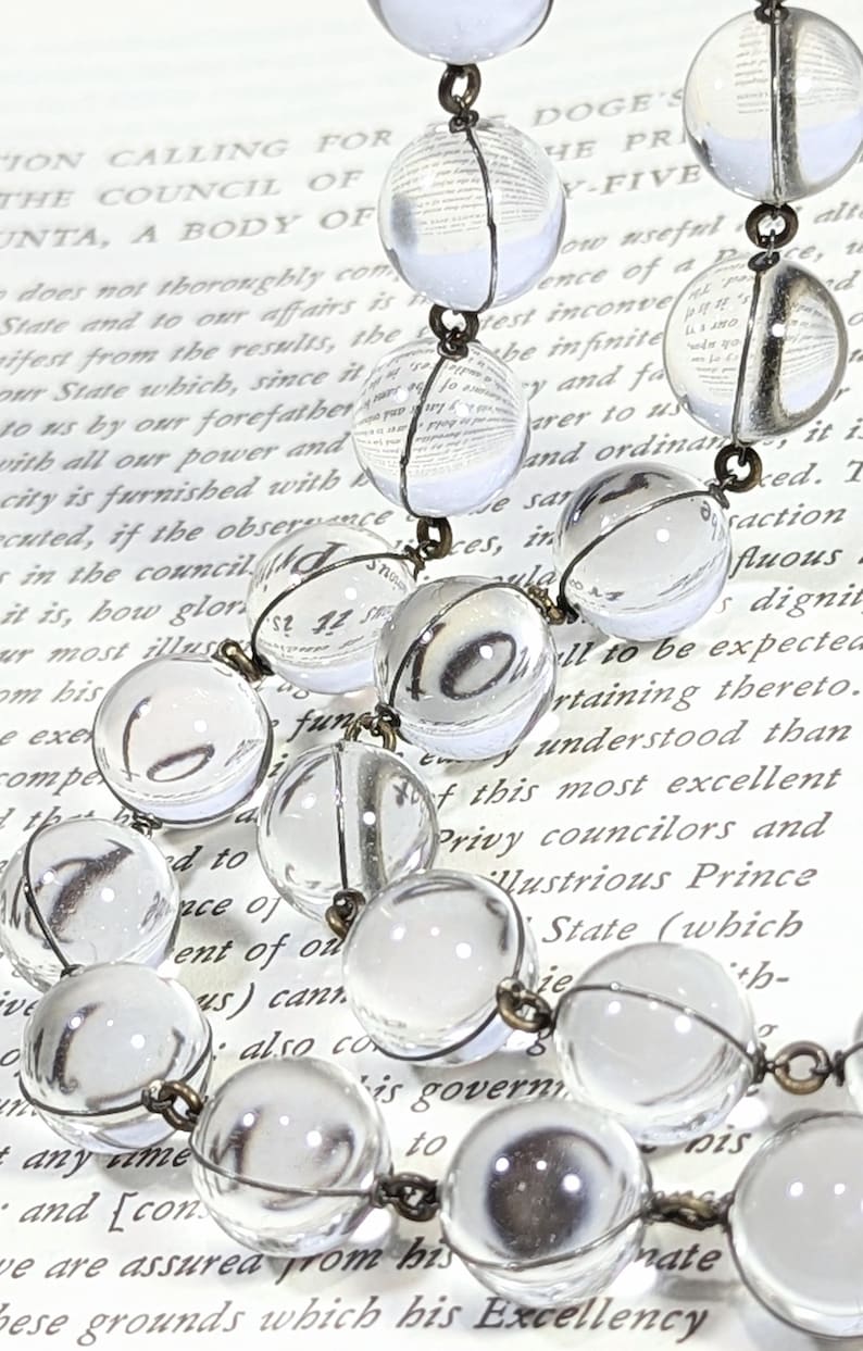 May include: A vintage glass bead necklace with clear glass beads and a silver wire. The beads are strung on a silver chain and the necklace is lying on a page of a book.