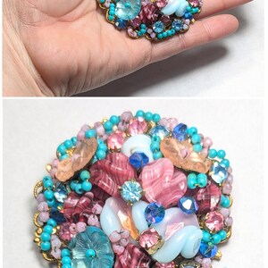 May include: A large, round brooch made of pink, blue, and white glass beads and rhinestones. The brooch is about 2 inches in diameter and is surrounded by a gold-colored metal frame.