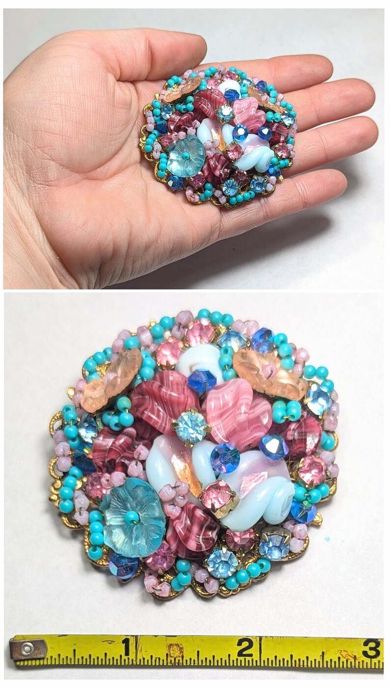 May include: A large, round brooch made of pink, blue, and white glass beads and rhinestones. The brooch is about 2 inches in diameter and is surrounded by a gold-colored metal frame.