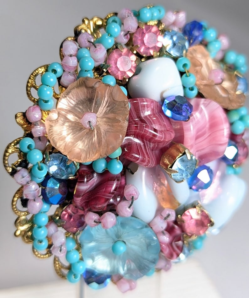 May include: A vintage brooch with a cluster of pink, blue, and white glass beads, flowers, and rhinestones. The brooch is set in a gold tone metal frame.