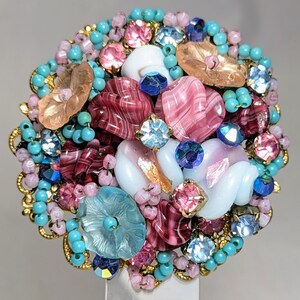 May include: A colorful brooch made of glass and beads. The brooch features pink, blue, and white glass pieces, as well as gold beads. The brooch is a circular shape and is approximately 2 inches in diameter.