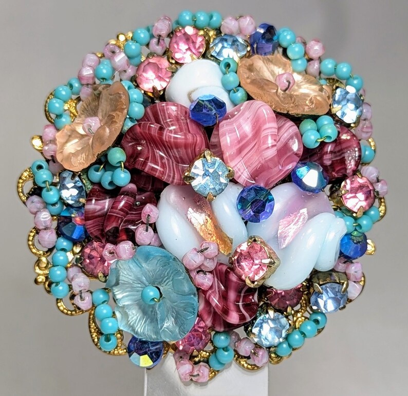 May include: A colorful brooch made of glass and beads. The brooch features pink, blue, and white glass pieces, as well as gold beads. The brooch is a circular shape and is approximately 2 inches in diameter.