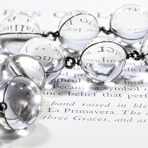 May include: A clear glass beaded necklace with a silver chain. The beads are round and reflect the light. The necklace is lying on a piece of paper with text.