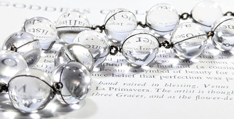 May include: A clear glass beaded necklace with a silver chain. The beads are round and reflect the light. The necklace is lying on a piece of paper with text.