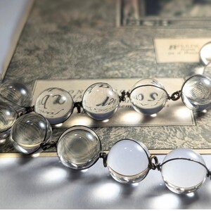 May include: A clear glass beaded necklace with a silver wire connecting the beads. The necklace is lying on a piece of paper with writing on it.