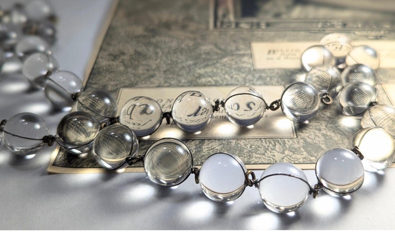May include: A clear glass beaded necklace with a silver wire connecting the beads. The necklace is lying on a piece of paper with writing on it.