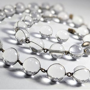 May include: A clear crystal beaded necklace with silver wire connecting the beads. The necklace is laid out in a loose circle on a white surface.