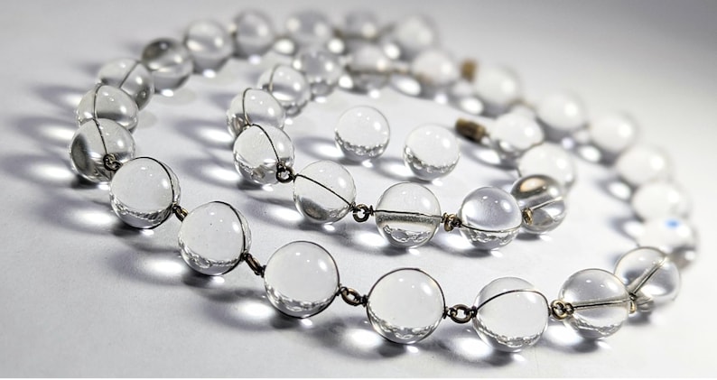 May include: A clear crystal beaded necklace with silver wire connecting the beads. The necklace is laid out in a loose circle on a white surface.