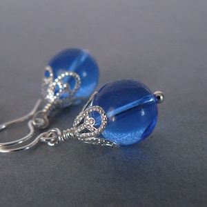 May include: A pair of earrings featuring round, translucent blue glass beads. Each bead is set within a decorative silver-toned metal filigree cap and attached to a simple silver-toned metal hook wire for pierced ears.