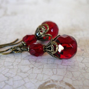 Puede incluir: Un par de pendientes con cuentas de cristal rojo en forma de lágrima y detalles de metal dorado adornados.