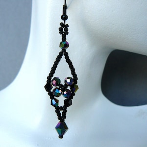 May include: A long dangle earring featuring a black wire hook and a diamond-shaped frame constructed from small matte black seed beads. The center and bottom of the earring are accented with faceted, iridescent dark purple and blue crystal beads.