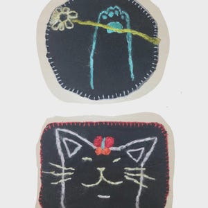 May include: Two embroidered fabric patches. The top circular black patch features a light blue cat paw holding a yellow-stemmed white flower. The bottom rectangular black patch displays a white-outlined cat face with a small orange and red butterfly.