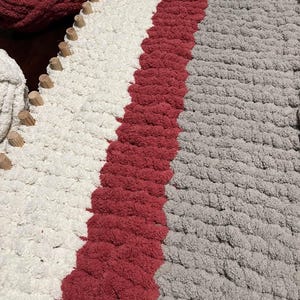 May include: A close-up of a knitted rug with three stripes of different colors: white, red, and gray. The rug is made with thick yarn and has a textured surface.
