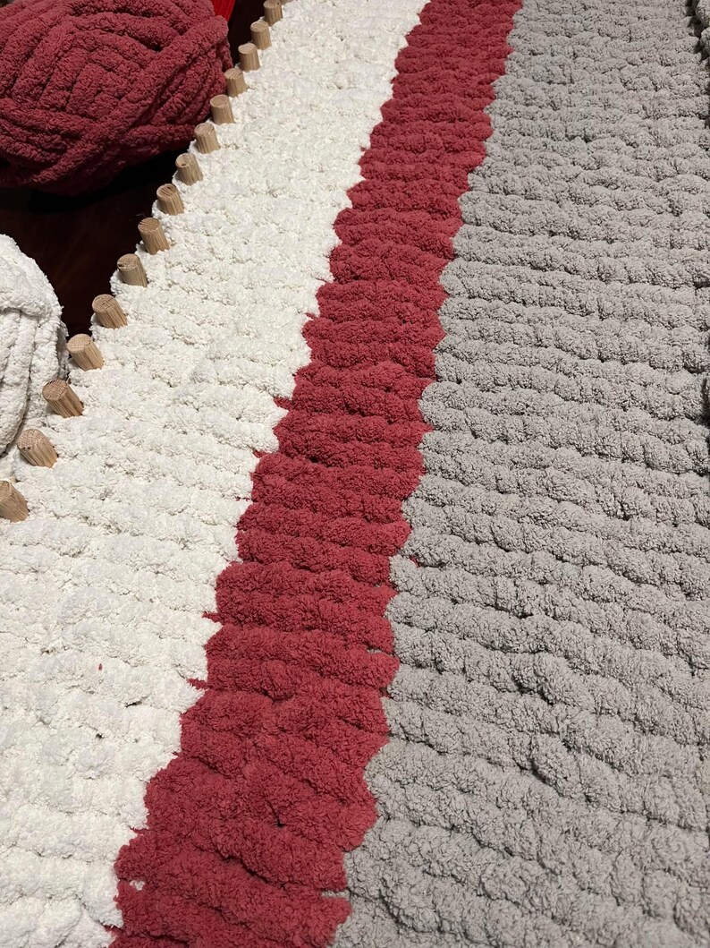 May include: A close-up of a knitted rug with three stripes of different colors: white, red, and gray. The rug is made with thick yarn and has a textured surface.