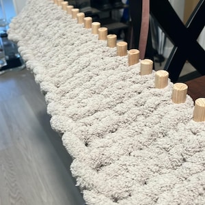 May include: A close-up of a beige knitted rug with wooden dowels along the edge. The rug is being made on a loom.