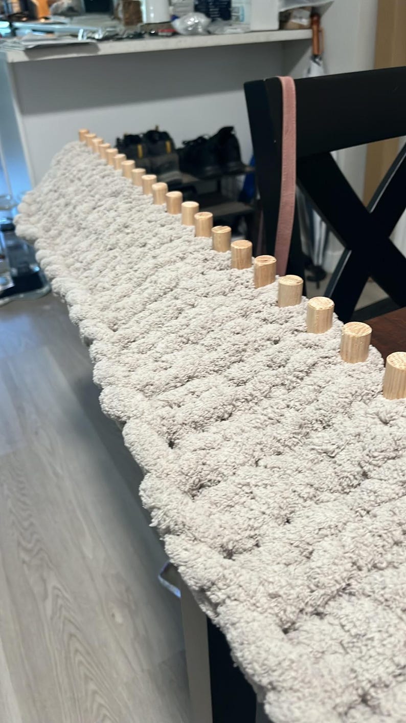 May include: A close-up of a beige knitted rug with wooden dowels along the edge. The rug is being made on a loom.