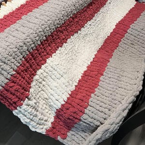 May include: A striped knitted blanket with pink, white, and gray yarn. The blanket is folded and draped over a chair.