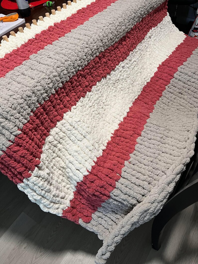 May include: A striped knitted blanket with pink, white, and gray yarn. The blanket is folded and draped over a chair.