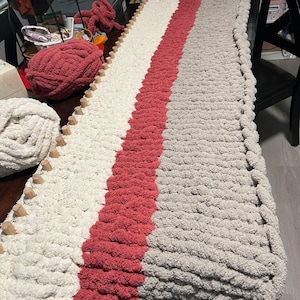 May include: A long, knitted throw blanket with a striped pattern in white, light pink, and gray. The blanket is being worked on with knitting needles and yarn.