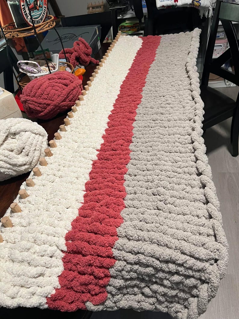 May include: A long, knitted throw blanket with a striped pattern in white, light pink, and gray. The blanket is being worked on with knitting needles and yarn.