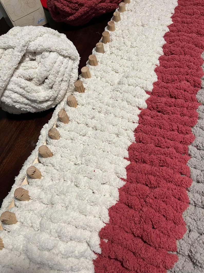May include: A close-up of a loom with white, red, and gray yarn being woven. The loom is made of wood and has dowels for holding the yarn in place.