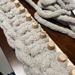 May include: A close-up of a gray yarn being woven onto a wooden loom with dowel rods. The yarn is fluffy and soft, and the loom is made of natural wood.