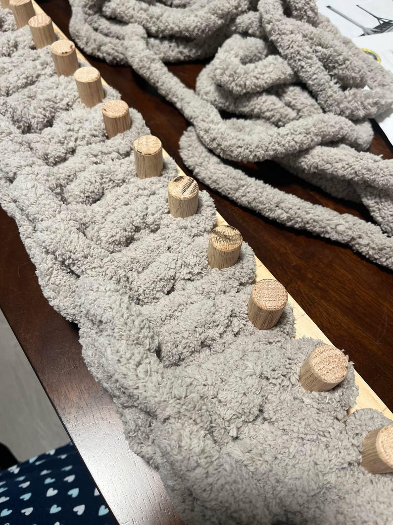 May include: A close-up of a gray yarn being woven onto a wooden loom with dowel rods. The yarn is fluffy and soft, and the loom is made of natural wood.
