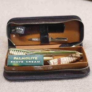 May include: Vintage toiletry kit with a black leather exterior and a brown interior. The kit contains a comb, razor, brush, and a box of Palmolive shave cream. The kit is open, revealing the contents.