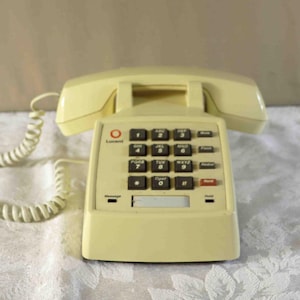 Könnte beinhalten: Cremefarbenes Vintage-Telefon mit einer Tastatur mit Druckknöpfen. Das Telefon hat eine Hörerablage und ein Spiralkabel. Die Tastatur hat schwarze Tasten mit weißen Zahlen und eine rote Taste. Das Telefon steht auf einer weißen Oberfläche mit Blumenmuster.