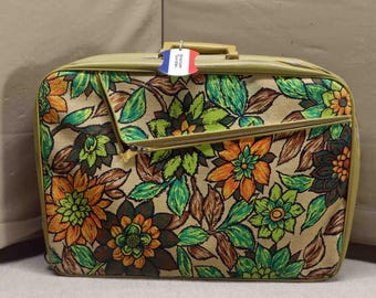 Puede incluir: Una maleta vintage con un estampado floral en tonos naranja, verde y marrón sobre un fondo beige. La maleta tiene un asa y un bolsillo con cremallera. Una etiqueta con los colores de la bandera francesa está adjunta.