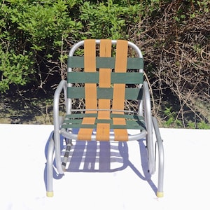 May include: A small outdoor chair featuring a silver-toned metal frame and a woven seat and backrest. The webbing consists of alternating vertical and horizontal straps in solid tan and dark green colors, creating a simple grid pattern.
