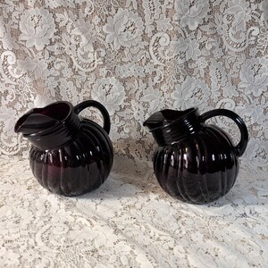 May include: Two matching dark purple glass pitchers featuring a rounded, ribbed body design with a smooth, curved handle and a distinct pouring spout. The glass has a deep, translucent plum hue and a glossy finish, reflecting light on the ridges.