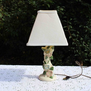 May include: A small, cream-colored table lamp with a square lampshade. The base is decorated with pastel-colored floral accents. The lamp has a brown electrical cord and plug. The lamp is on a white, textured surface.