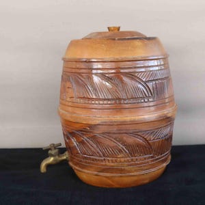 Puede incluir: Un dispensador de agua de madera marrón en forma de barril con tapa y grifo de latón. El barril presenta patrones tallados en forma de hojas alrededor del centro y la parte inferior. El dispensador está diseñado para contener y dispensar líquidos.