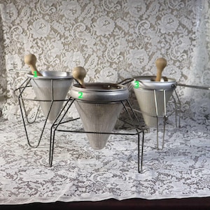 May include: Three silver-coloured metal conical food strainers, each featuring a fine mesh body, a sturdy wire stand, and a wooden pestle handle. The strainers are numbered one, two, and three in green, and each includes a long metal handle for support.