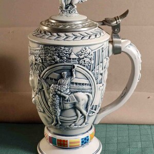 Puede incluir: Una jarra de cerveza de cer&aacute;mica blanca y azul con un dise&ntilde;o en relieve detallado que representa una escena de carreras de caballos. La tapa presenta una figura de caballo y jinete. La base tiene una banda colorida de patrones geom&eacute;tricos. El asa es ornamentada.