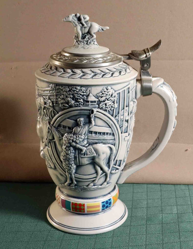 Puede incluir: Una jarra de cerveza de cer&aacute;mica blanca y azul con un dise&ntilde;o en relieve detallado que representa una escena de carreras de caballos. La tapa presenta una figura de caballo y jinete. La base tiene una banda colorida de patrones geom&eacute;tricos. El asa es ornamentada.