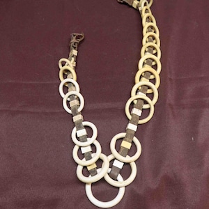 May include: A necklace featuring a series of off-white circular links connected by brown leather and metal accents. The necklace has a clasp at each end and is displayed against a dark burgundy background.